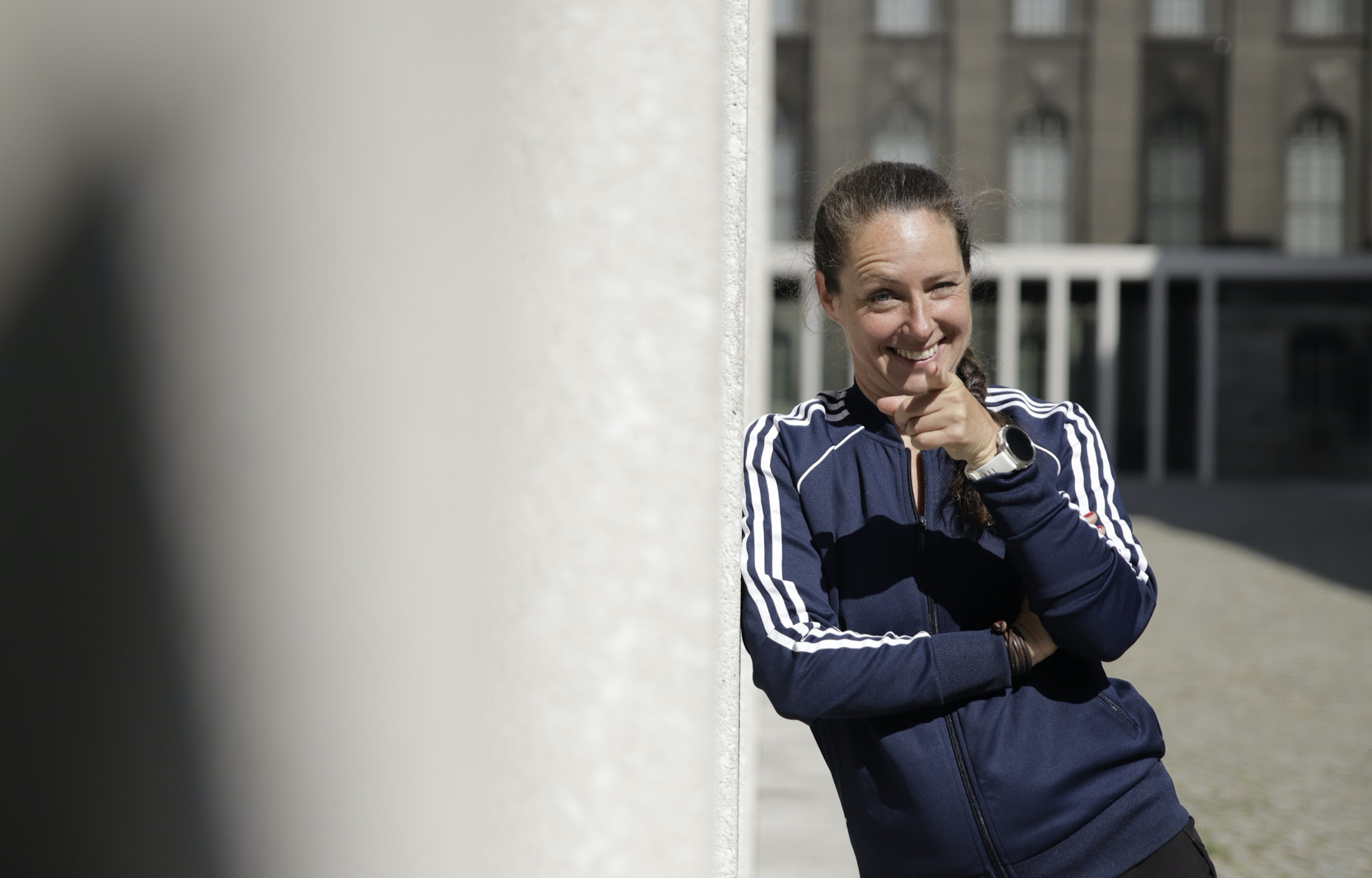 Eine lächelnde Frau in einem blauen Trainingsanzug lehnt an einer Wand und zeigt mit dem Finger auf sich.
